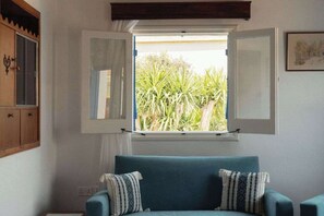 A room with a teal sofa, a window with open shutters, and a view of greenery outside. - Charming 2BR Village Getaway Near the Beach (Agios Theodoros Larnakas)