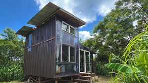 A two-story wooden cabin with a glass balcony, surrounded by greenery and a clear sky.