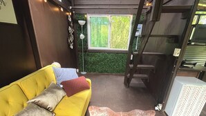 A yellow sofa with patterned cushions, a large brown leather rug, a green wall with a window, and a ladder.