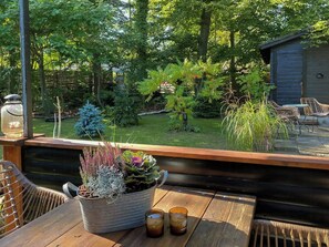 Eine Holzterrasse mit Tisch, Stühlen, einem Blumentopf und Blick auf einen Garten mit Bäumen und einem Gartenhaus.