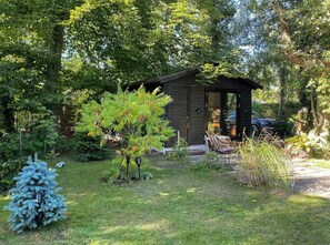 A wooden cabin with a covered porch, surrounded by lush greenery and a small tree. - House in Skæring near city and nature (Egå)