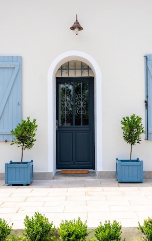 Ein Gebäude mit einer dunkelblauen Tür, weißen Wänden und blauen Fensterläden. Vor der Tür befinden sich je ein Blumentopf auf beiden Seiten.