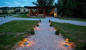 Un sendero de grava conduce a una estructura de piedra con un pérgola, rodeada de árboles pequeños y un césped.