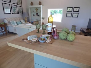 A kitchen counter with various food items, a bottle of olive oil, and a dining area with a sofa and a window. - Latour - Spacious elegance in the grounds of Chateau Mirabel (Puylaurens)