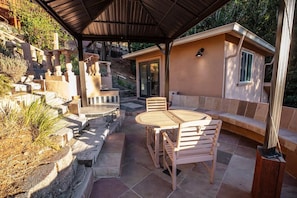Een buitenterras met een houten tafel en stoelen, een stenen openhaard en een overdekte zithoek.