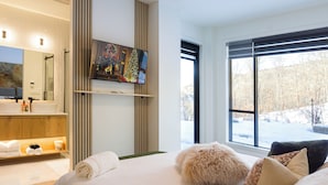 A modern hotel room with a large bed, a television mounted on the wall, a bathroom with a mirror and sink, and a window with a view of the outdoors.