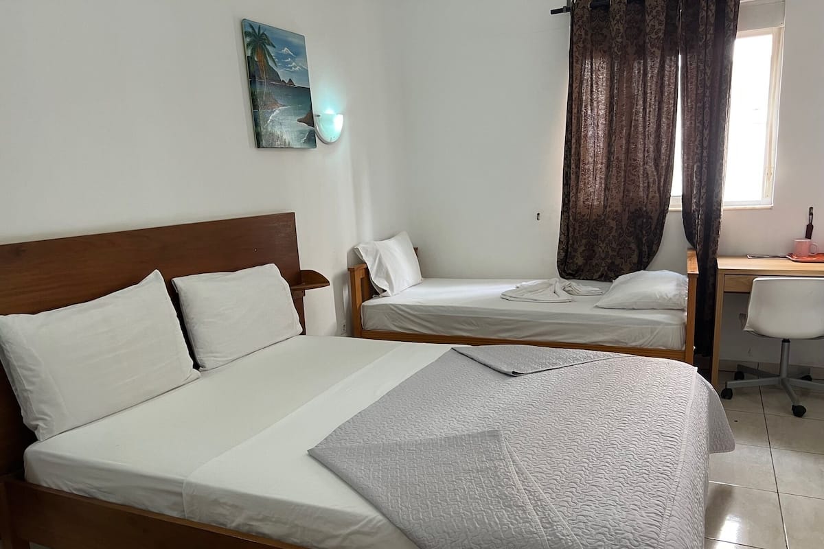 A hotel room with two single beds, a desk with a chair, a window with curtains, and a wall-mounted light fixture.
