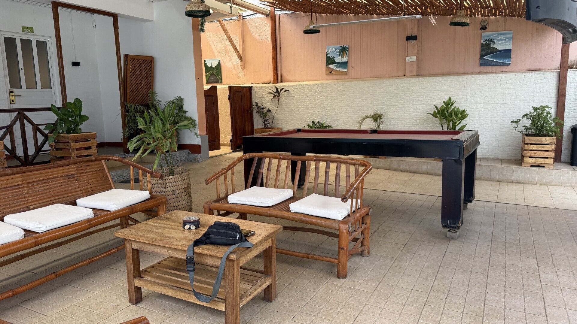 A pool table, wooden chairs with cushions, a small table with a bag, and potted plants in a room with a tiled floor and a wooden ceiling.