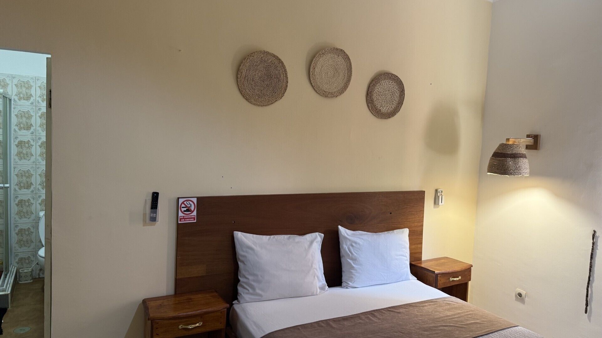 A hotel room with a wooden headboard, two white pillows, a bed with a brown blanket, a nightstand with a drawer, a wall-mounted lamp, and a no-smoking sign.