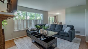 Een moderne woonkamer met een donkergrijze bank, een houten salontafel en een groot raam met witte luiken.