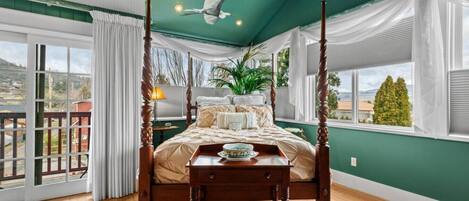 A four-poster bed with a wooden headboard, a ceiling fan, and a view of the outside through large windows.