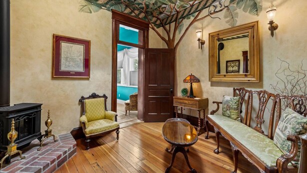 Une pièce avec une cheminée, un plancher en bois, un fauteuil vert, un banc en bois, un miroir et un mur orné d’une fresque représentant un arbre.
