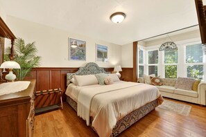 A bedroom with a bed, a nightstand, a dresser, a window with blinds, and a view of greenery outside.