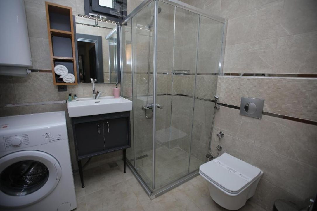 A modern bathroom with a glass-enclosed shower, a white sink, a washing machine, and a toilet.