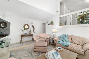 Una sala de estar moderna con un sofá, un reposapiés y una mesa de centro. Hay una mesita de noche con una lámpara y un espejo en la pared.