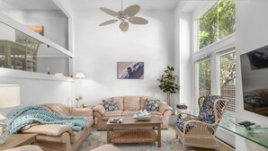 Ein geräumiges Wohnzimmer mit einem beigefarbenen Sofa, einem Couchtisch, einem Korbstuhl und einem großen Fenster mit Jalousien.