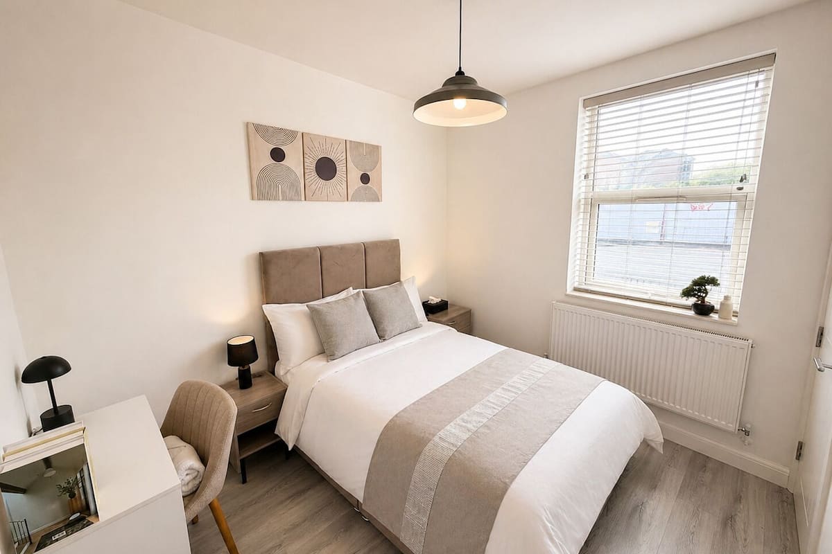 A bedroom with a bed, bedside table, lamp, chair, artwork, and a window with blinds.