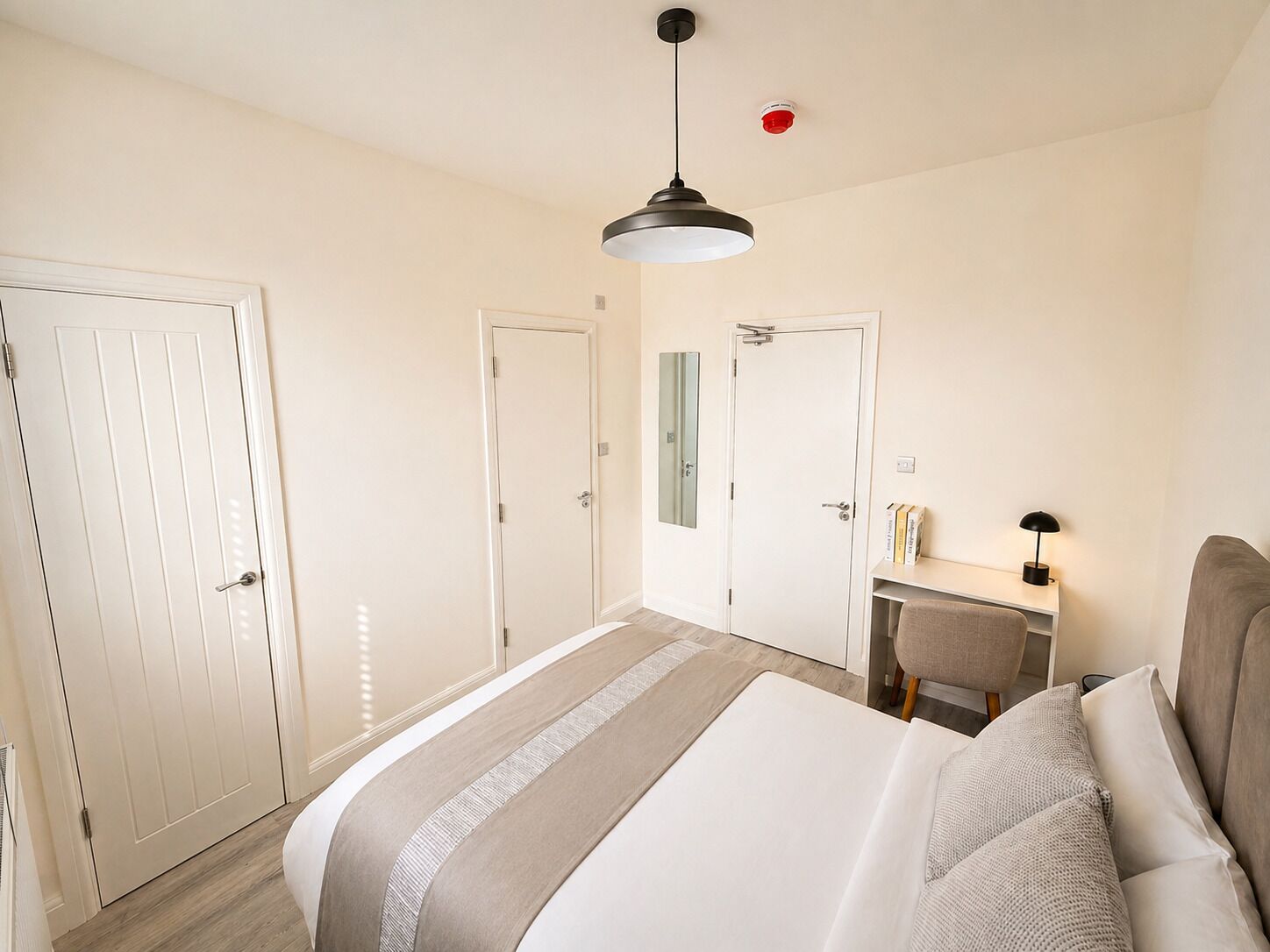 A bedroom with a bed, a desk, a chair, a hanging light fixture, and a door.