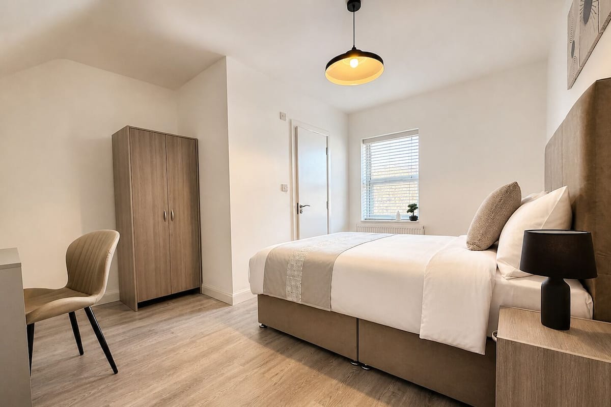 A bedroom with a bed, a chair, a wardrobe, a bedside table with a lamp, a window with blinds, and a hanging light fixture.