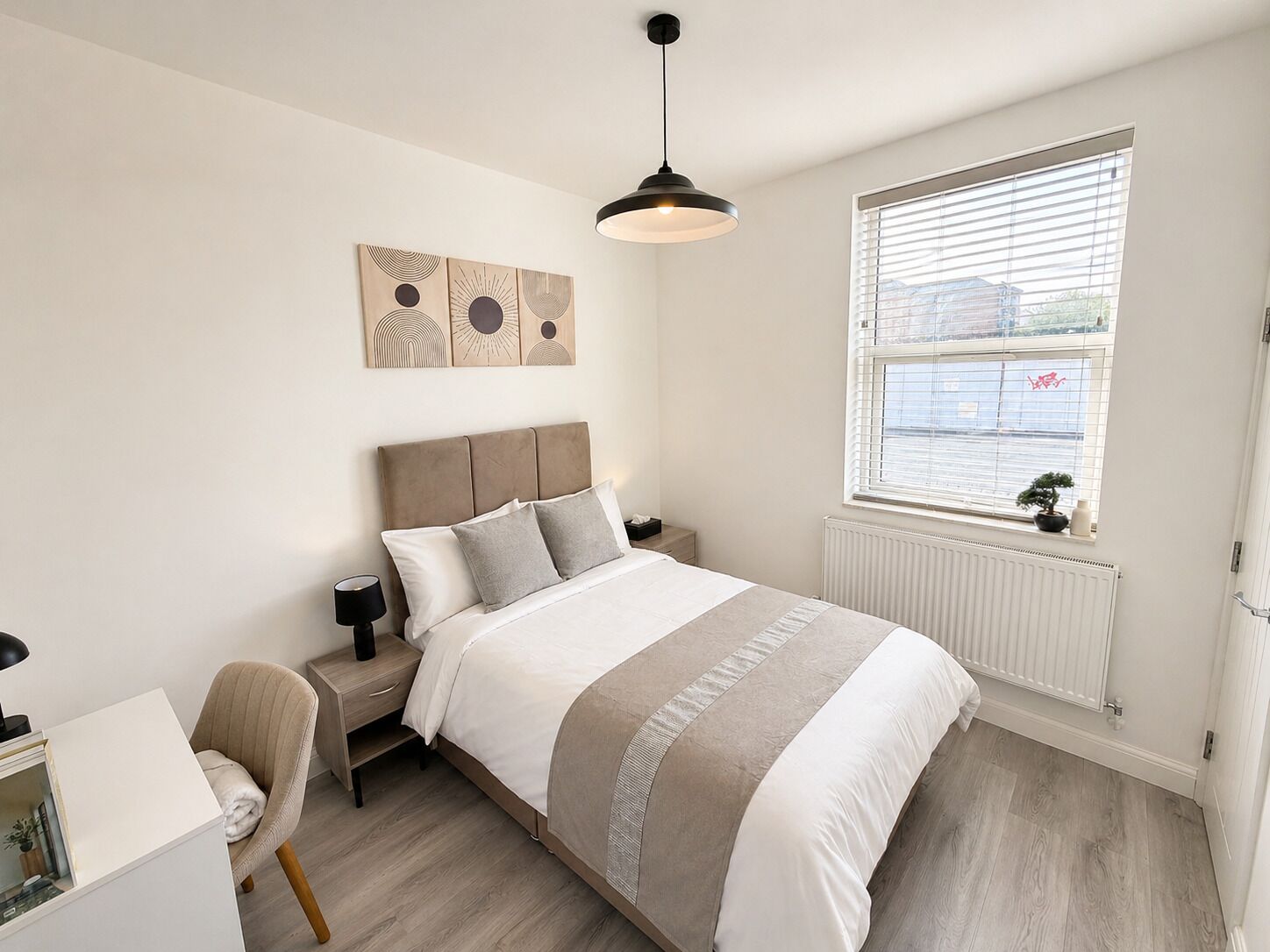 Une chambre à coucher comprenant un lit, une table de chevet, une chaise, une œuvre d’art, une fenêtre et un radiateur.