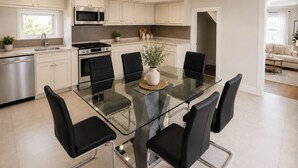 A modern kitchen with a glass dining table, black chairs, stainless steel appliances, and light-colored cabinetry. - New England Coastal Residence (Gloucester)
