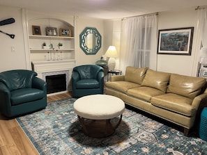 A living room with a fireplace, two leather armchairs, a sofa, a round ottoman, a patterned rug, a mirror, a lamp, and a painting on the wall.