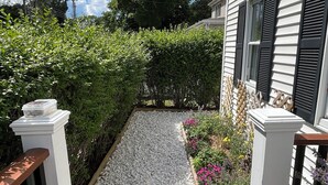 A pathway leading to a house with a white exterior and black shutters, surrounded by greenery and a gravel area. - New England Coastal Residence (Gloucester)