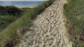 Ein sandiger Pfad führt zu einem Strand, begleitet von grasbewachsenen Dünen.