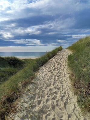 Ein sandiger Pfad führt zu einem Strand, begleitet von grasbewachsenen Dünen.