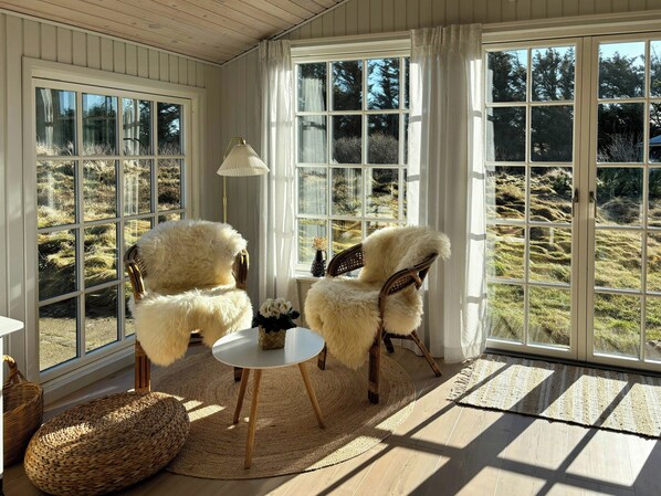Ein Zimmer mit zwei bequemen Sesseln, einem kleinen runden Tisch, einer Lampe und Blick ins Freie durch große Fenster.