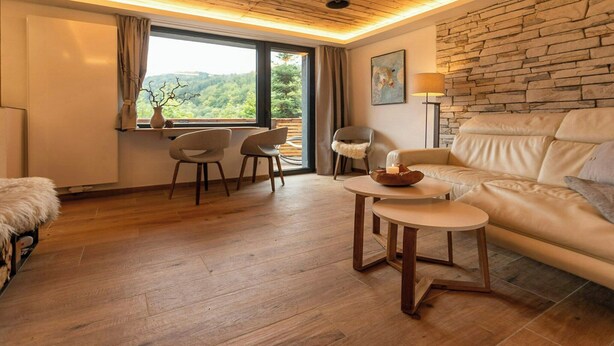 Ein Zimmer mit Holzboden, Steinwand, einer Couch, einem Tisch und einem Fenster mit Blick ins Grüne.