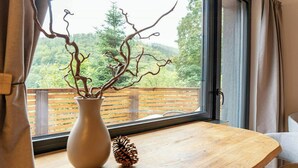 Ein Holztisch mit einer Vase und einer Kiefernzapfe neben einem Fenster mit Blick auf eine Holzterrasse und Bäume.