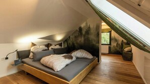 Ein Schlafzimmer im Loft-Stil mit schrägem Dach, einem Bett mit grauer Bettwäsche und einer Wandmalerei von einem Wald.