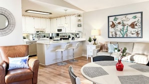 Un salon moderne comprenant un coin repas, une cuisine avec des meubles blancs et une grande œuvre d’art décorative représentant des poissons, qui orne un mur.