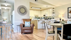 Une cuisine moderne avec des meubles blancs, un îlot central et un coin repas comprenant un fauteuil en cuir marron et des chaises blanches.