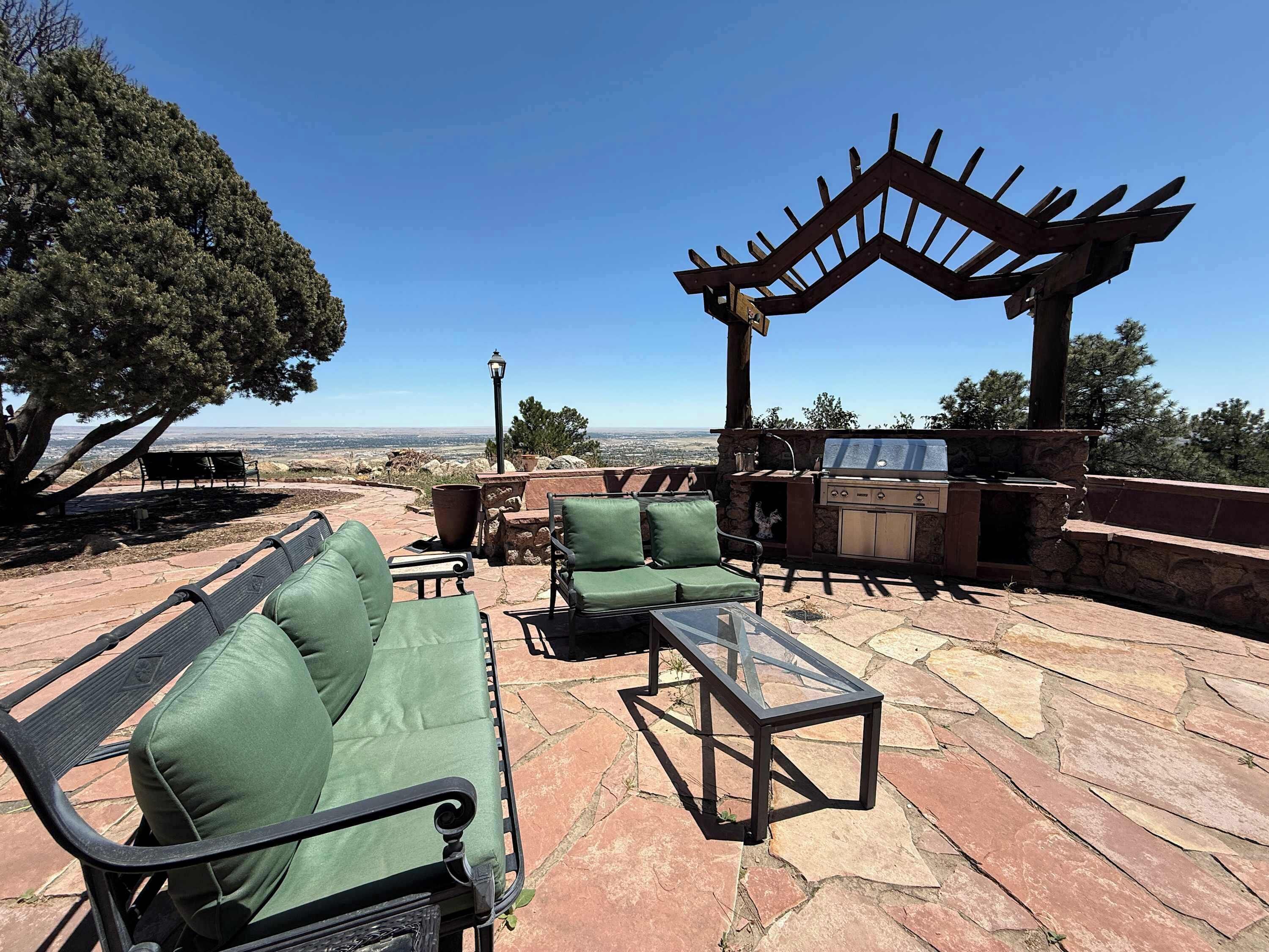 Eine Terrasse im Freien mit Steinböden, grünen Sitzmöbeln, einem Tisch mit Glasplatte und einem Grill.