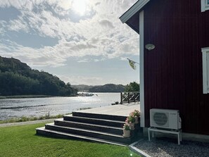 A house with a wooden deck, steps leading to a door, a flag, and a view of a lake. - New timber holiday home with bay views (Kungshamn)