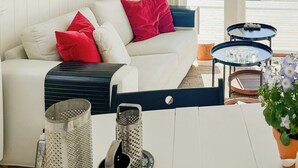 En hyggelig stue med en hvid sofa, røde puder, et lille bord med glas, en rivjern og en potteplante.