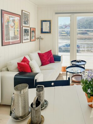 A cozy living room with a white sofa, red pillows, a small table with glasses, a grater, and a potted plant. - New timber holiday home with bay views (Kungshamn)