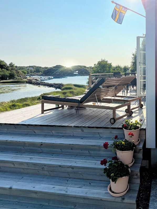 A wooden deck with lounge chairs, potted plants, and a view of a body of water with boats. - New timber holiday home with bay views (Kungshamn)