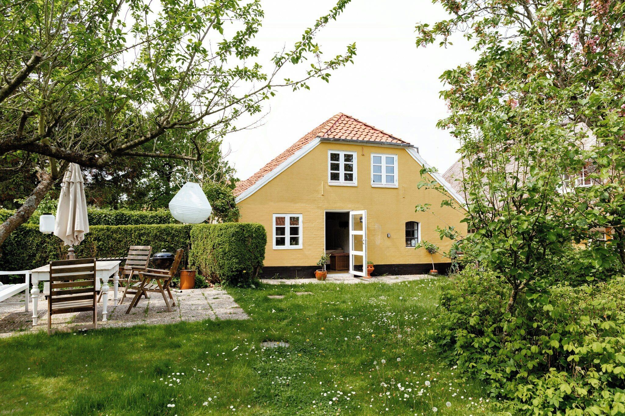 Ein gelbes Haus mit rotem Dach, einer weißen Tür und einem kleinen Garten mit Tisch und Stühlen.