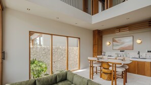 A modern living room with a sectional sofa, a dining area with a table and chairs, and a kitchen with wooden cabinets and a stone wall. - Cozy villa, prime Uluwatu, actual walking distance (Badung Regency)