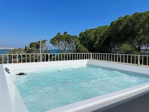 Ein Whirlpool mit weißer Außenverkleidung, umgeben von einem weißen Geländer, mit Blick auf ein Gewässer und Grünflächen.