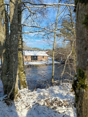 Ein rustikales Gebäude mit schneebedecktem Dach, umgeben von Bäumen und einem Gewässer.