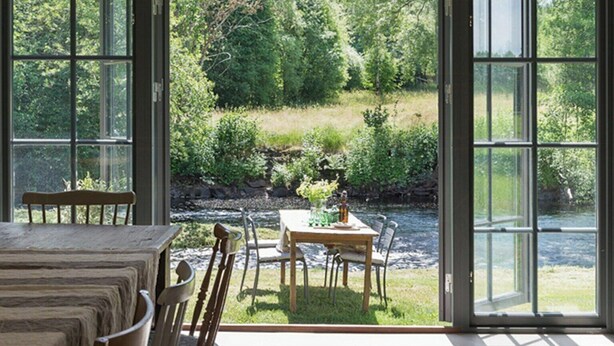 Un comedor con mesa y sillas, y vistas a un río y follaje a través de amplios ventanales.