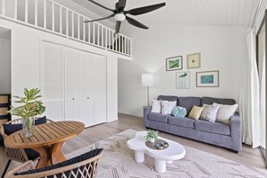 A room with a ceiling fan, a grey sofa, a wooden table, and a white lamp. - Turtle Bay, Golf Course Views Walk to Beach (Kahuku)