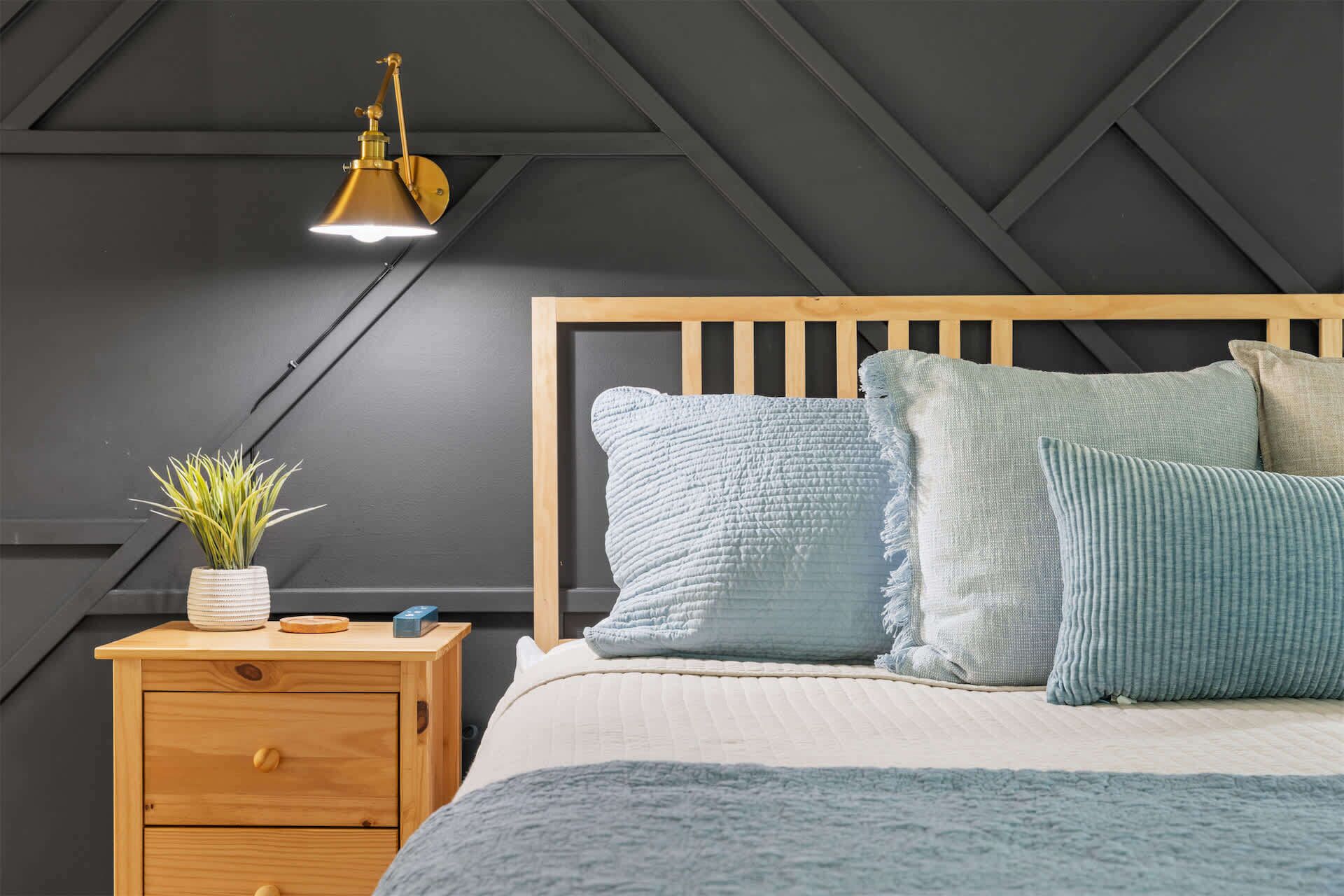 Une chambre à coucher avec une table de chevet en bois, une lampe de chevet, une plante en pot et un lit agrémenté de plusieurs oreillers.