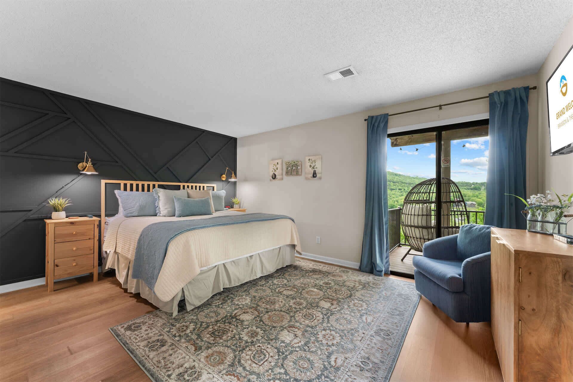 Une chambre à coucher comprenant un lit, des tables de chevet, un bureau, une chaise, une grande fenêtre avec des rideaux et une vue sur la verdure.