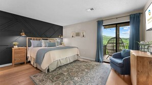 Een slaapkamer met een bed, nachtkastjes, een bureau, een stoel, een groot raam met gordijnen en uitzicht op groen.