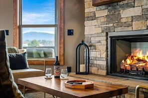 Ein gemütliches Wohnzimmer mit Kamin aus Stein, einer Couch, einem Couchtisch mit Gläsern und einer Flasche und Blick ins Freie.